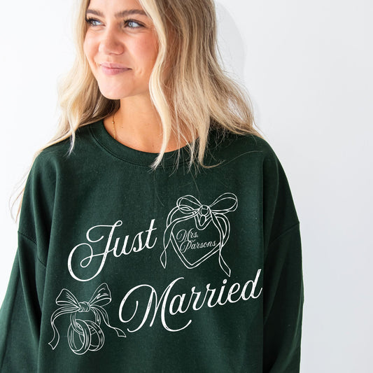 girl wearing a forest green crewneck sweatshirt with a custom just married design featuring a heart locket with a custom name in the center and wedding rings tied with a bow. design is printed large on the center chest in white ink