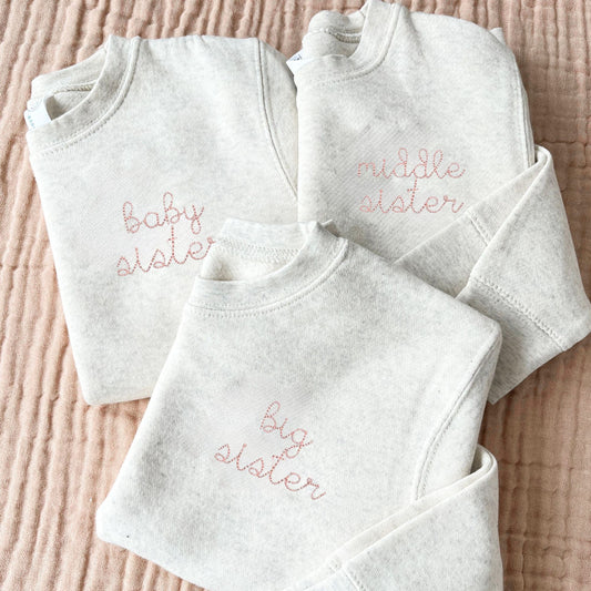 three natural heather toddler crews with big sister, middle sister, and baby sister stitched on the left chest in powder pink thread