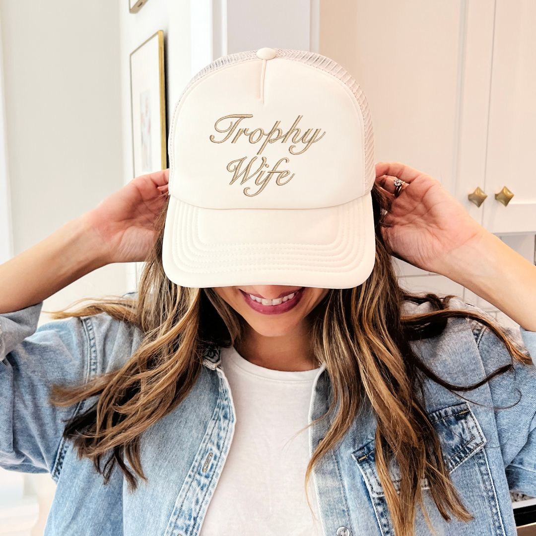 young woman wearing a sand trucker hat with embroidered Trophy Wife text in camel thread