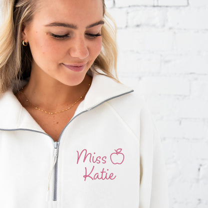 close up of woman wearing a white boxy ladies fit quarter zip pullover with a custom name and outlined apple embroidered in a pink thread.