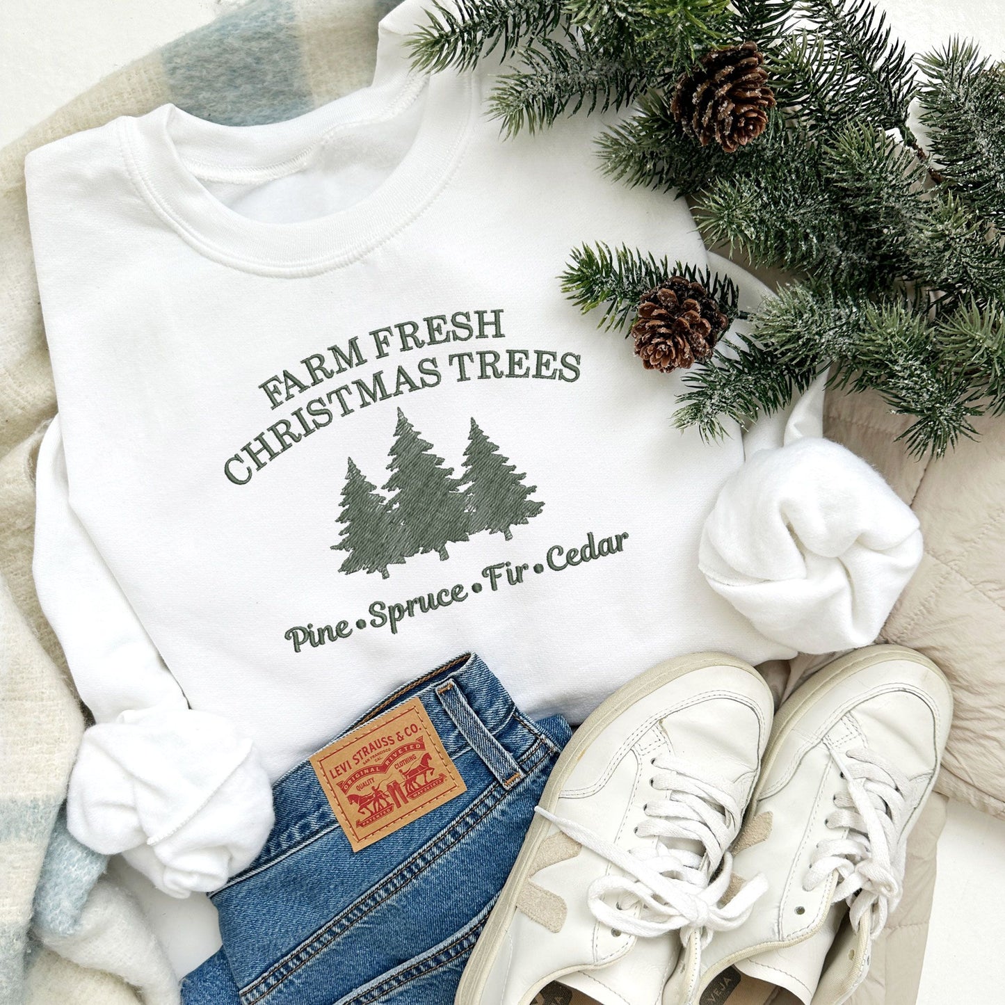 Christmas styled flat lay ff a white crewneck sweatshirt with embroidered farm fresh Christmas trees design with Christmas tree embroidery across the chest in ivy green thread