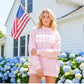 Woman wearing a pink sweatshirt with 'CUSTOM' text in front of a house with an American flag.