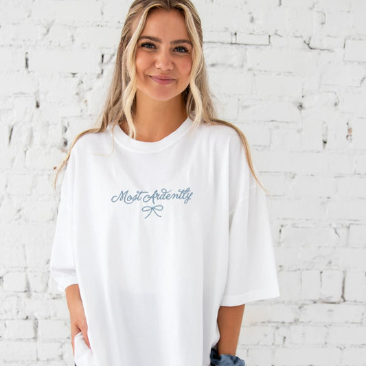 young woman wearing a white comfort colors t-shirt with embroidered most ardently and bow design in baby blue thread