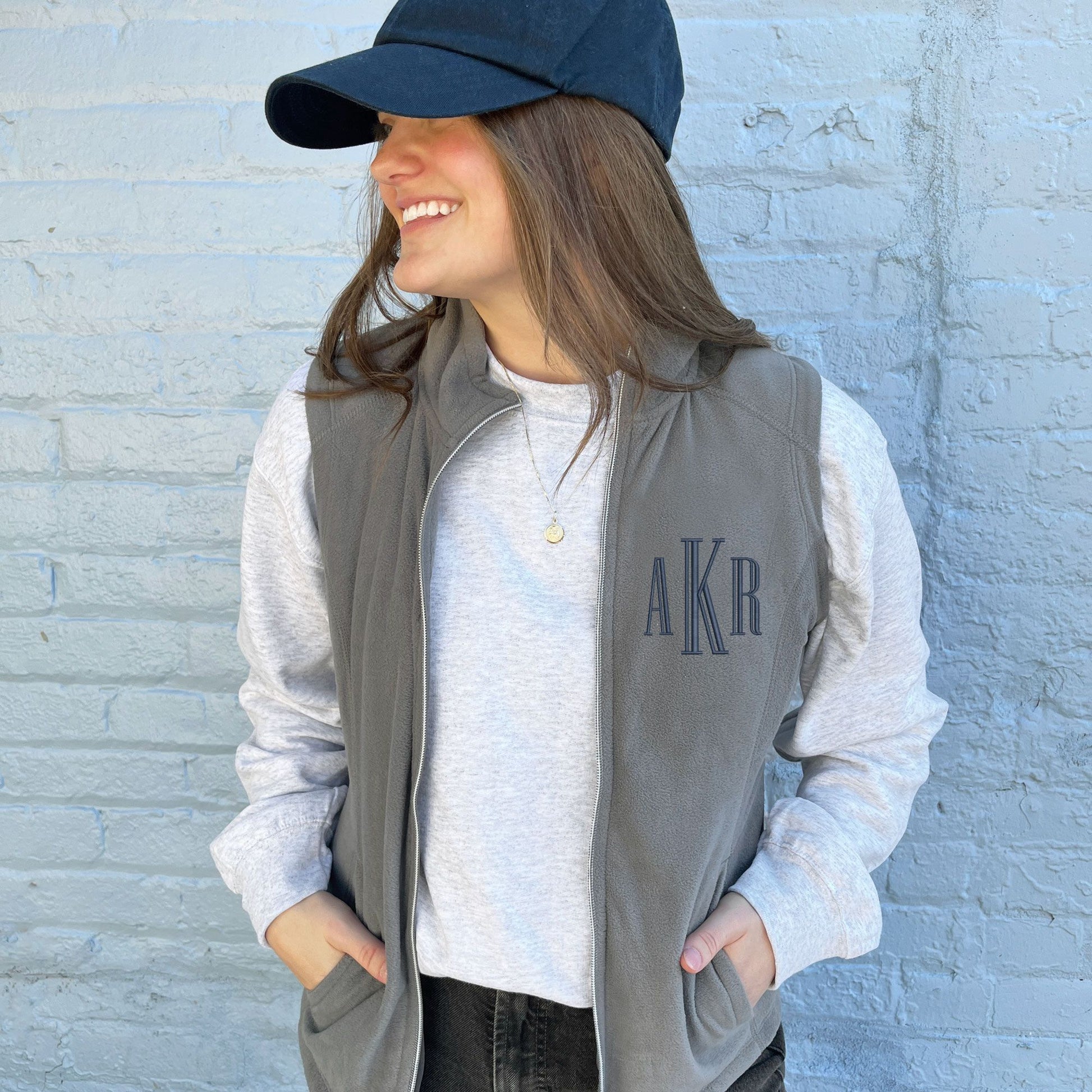 young woman wearing a lightweight grey fleece vest with a navy blue embroidered monogram on the left chest