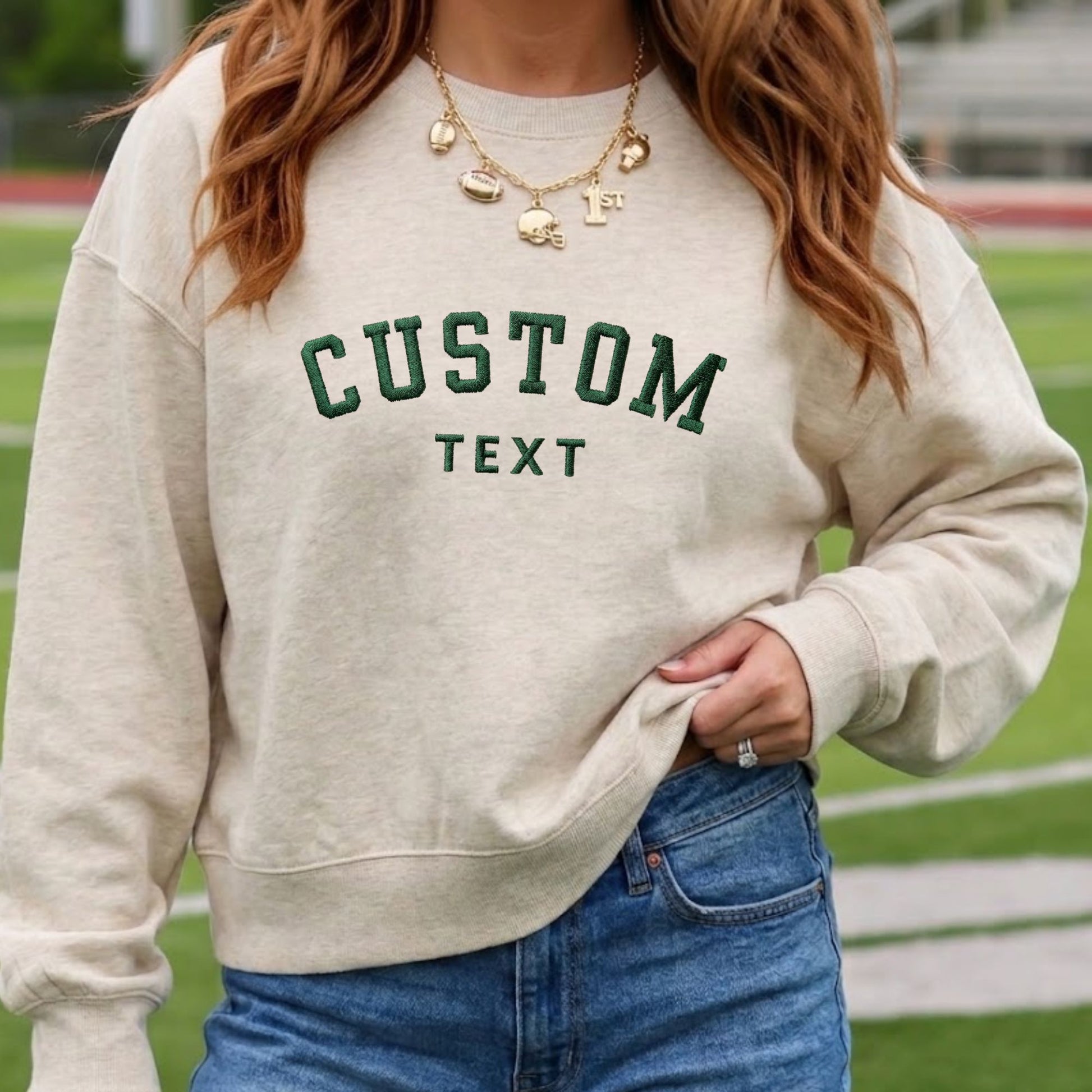 A woman wearing a cream-colored crewneck sweatshirt with 'CUSTOM TEXT' embroidered in dark green on the front, paired with blue jeans and gold necklaces, set against a blurred outdoor athletic field background.