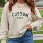 A woman wearing a cream-colored crewneck sweatshirt with 'CUSTOM TEXT' embroidered in dark green on the front, paired with blue jeans and gold necklaces, set against a blurred outdoor athletic field background.