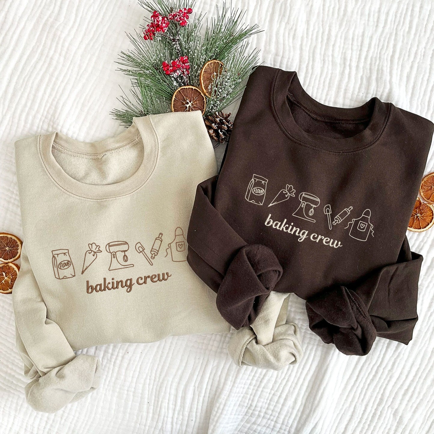 Christmas styled flat lay of two crewneck sweatshirts in sand and dark chocolate colors. embroidered on the center chest of the sweatshirts are outlined baking icons and baking crew text