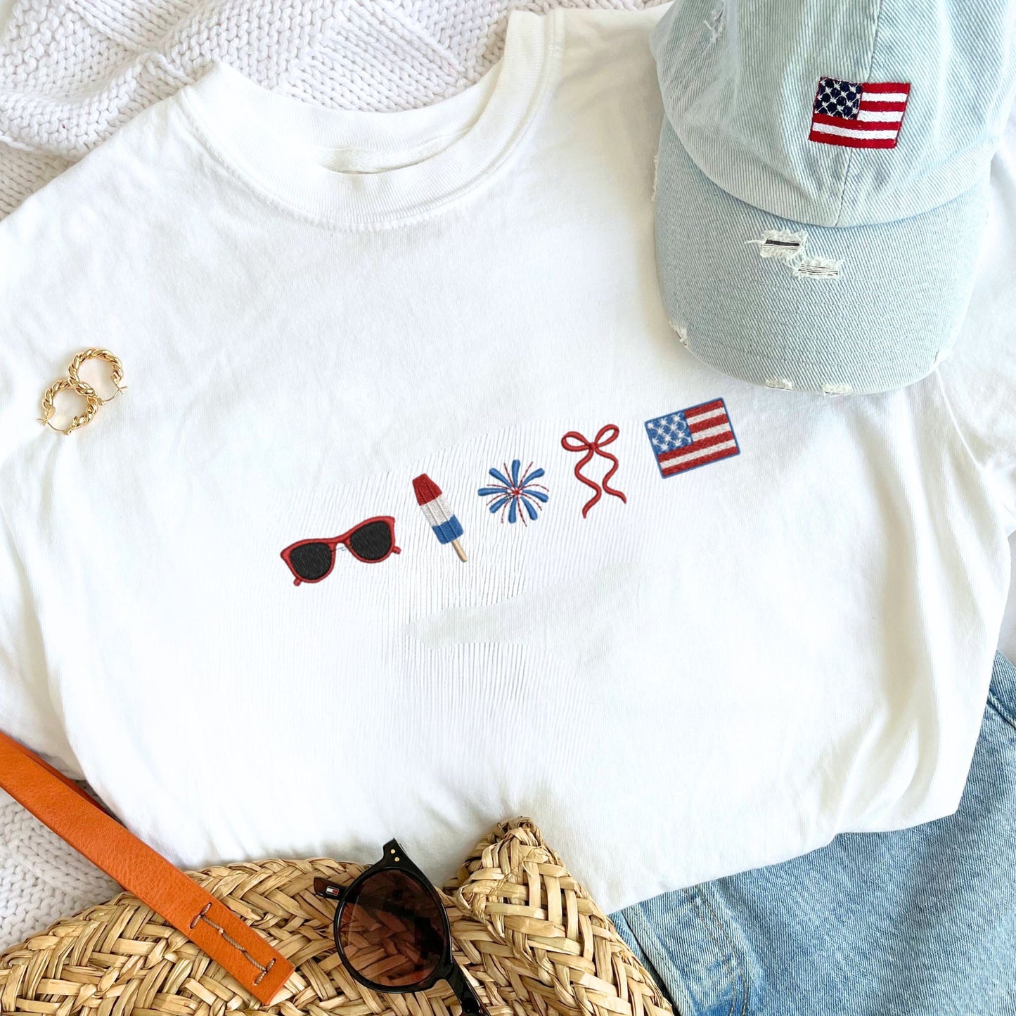 flat lay image of an outfit featuring a straw round tote, sunglasses, jean shorts, gold hoops, distressed denim hat with a mini American flag embroidery, and a white t-shirt with 4th of July inspired embroidered icons: red sunglasses, a red white and blue popsicle, firework, red bow, and an American flag.