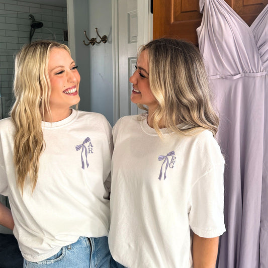 Two girls wearing white comfort colors t-shirts with custom ribbon bow and stacked initial embroidery on the left chest in smoky orchid thread.