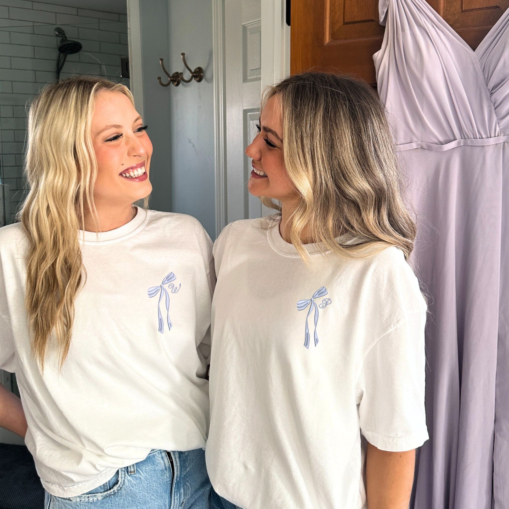 two girls wearing white comfort colors t-shirts with embroidered ribbon bow and cursive initial embroidered on the left chest in lilac thread