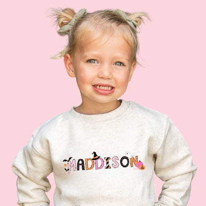 Little girl wearing a heather oat crewneck sweatshirt with a custom name print in mix of pink, orange, and black halloween patterned text decorated with cute spooky season icons.