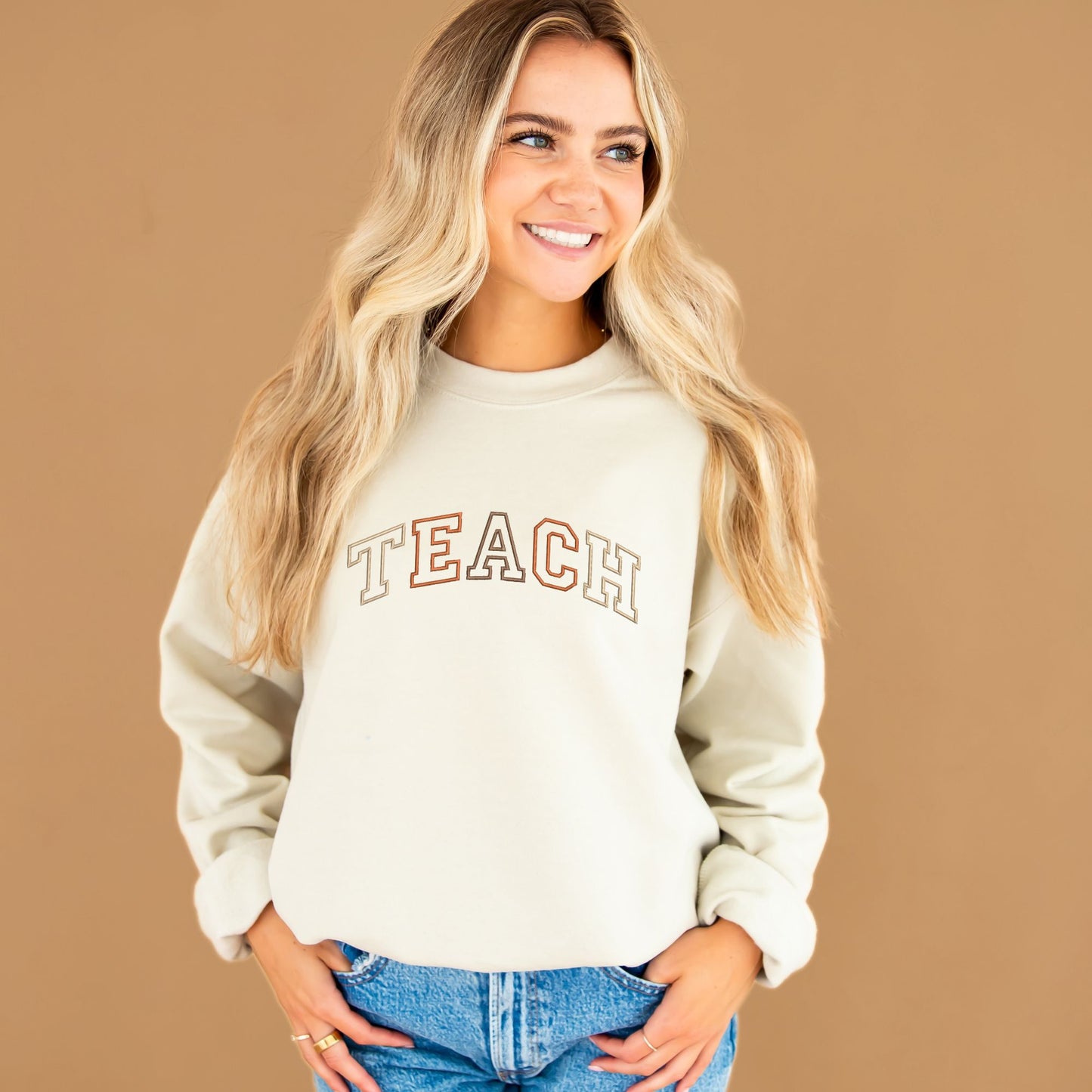 young woman wearing a sand crewneck sweatshirt with embroidered TEACH in fall colors across the chest
