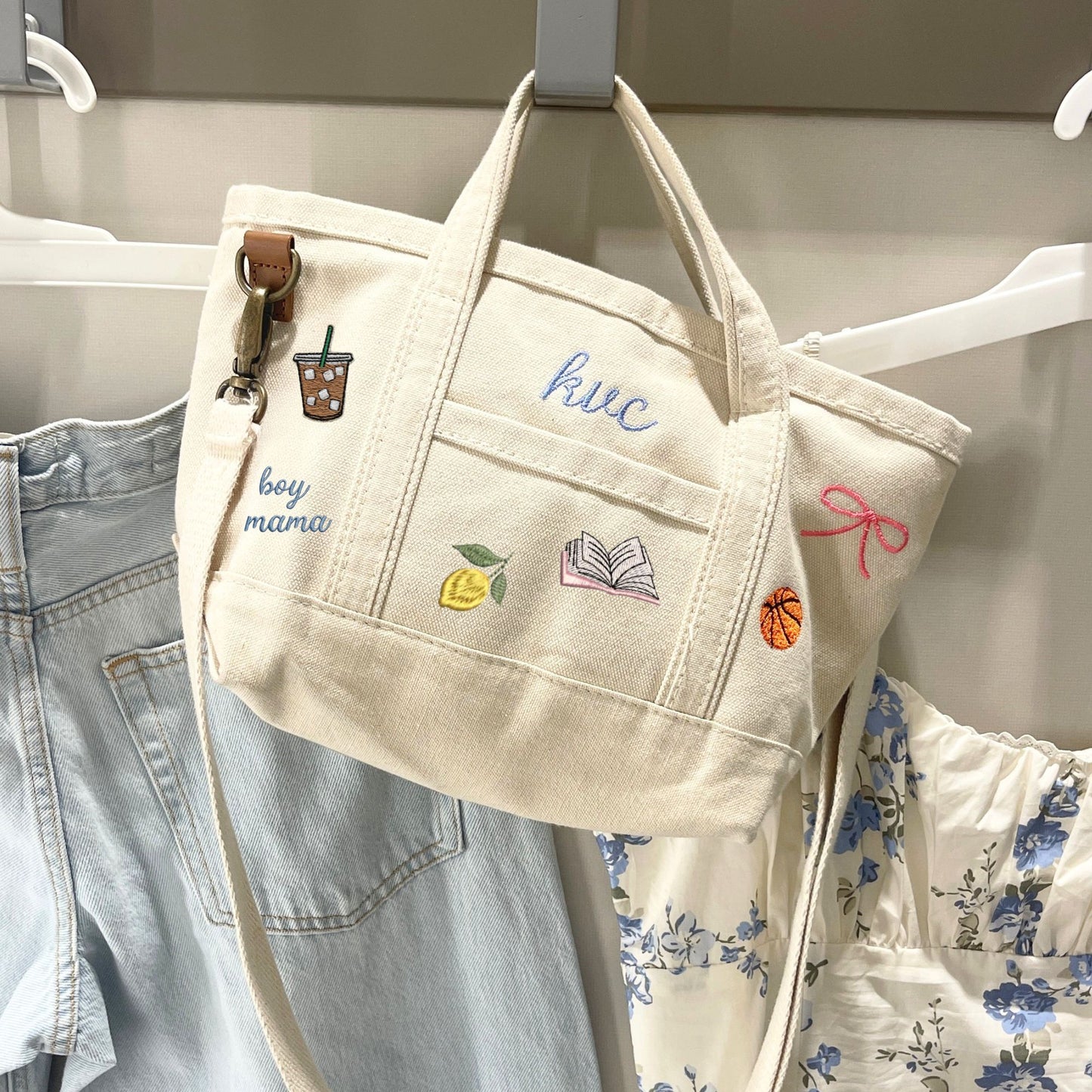Beige tote bag with embroidered designs hanging on a rack with clothes.