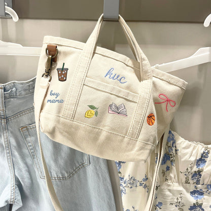 Beige tote bag with embroidered designs hanging on a rack with clothes.