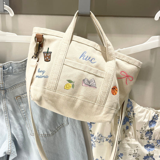 Beige tote bag with embroidered designs hanging on a rack with clothes.
