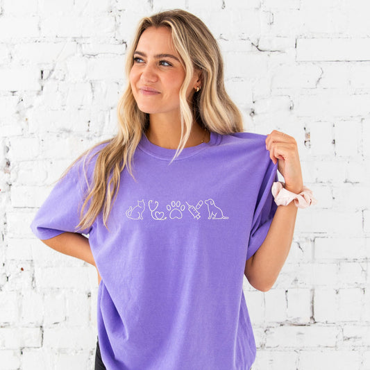 young woman wearing a violet tee with embroidered veterinarian icons across the chest in white thread.