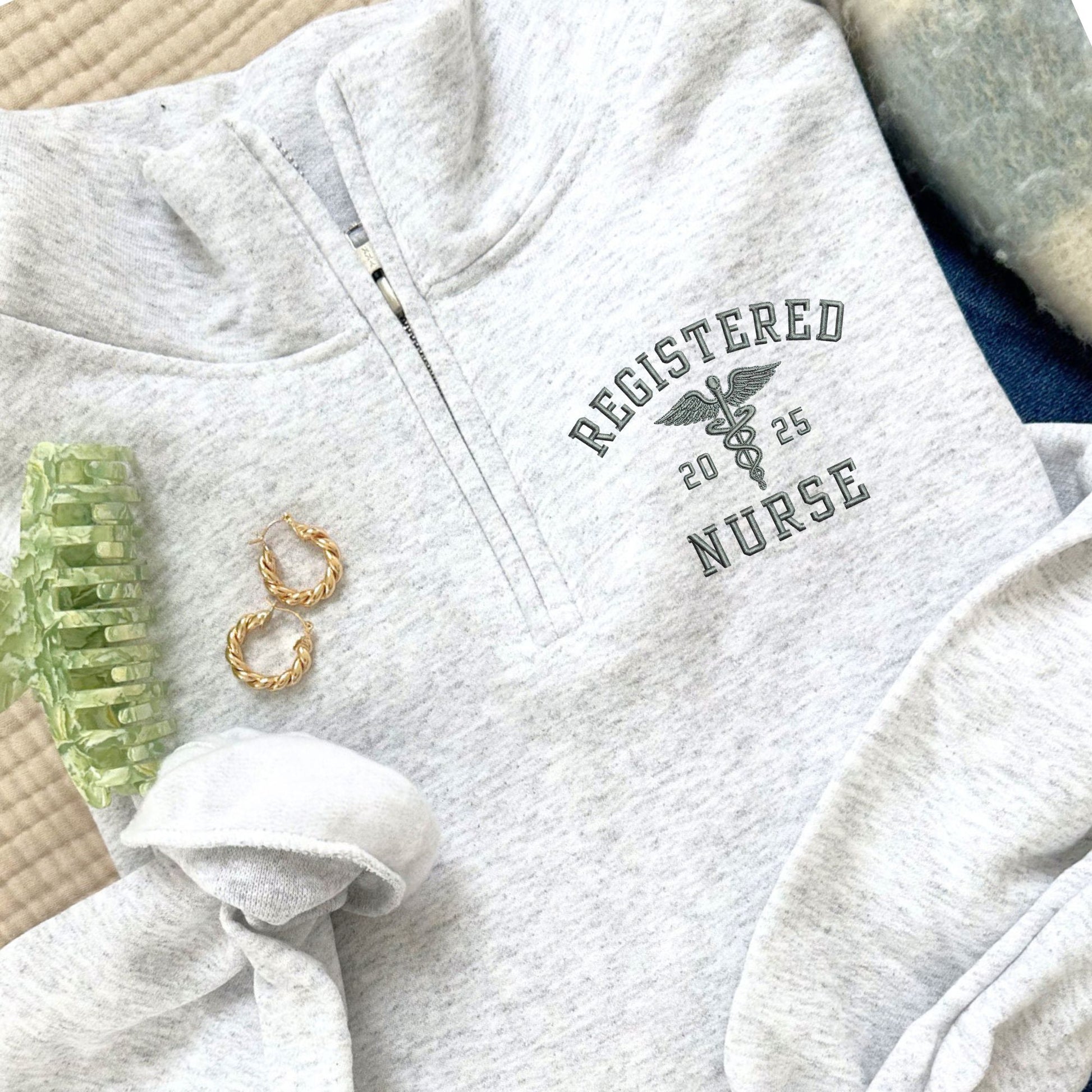 Gray sweatshirt with 'Registered Nurse 2025' text and caduceus symbol on a surface with accessories.