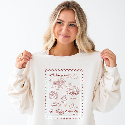A woman with blonde hair holds up a cream-colored crewneck sweatshirt with a rust graphic print on the front. The graphic features a stamp-like border with illustrations of a covered bridge, tree, pumpkins, and the text 'with love from.