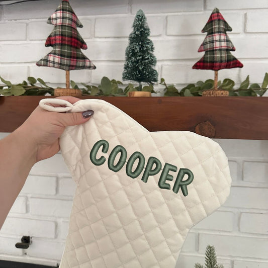 cream quilted Christmas stocking in the shape of a dog bone with embroidered name Cooper in ivy green near the top of the stocking