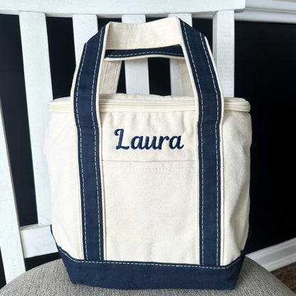 Beige tote bag with navy handles and trim, embroidered with 'Laura', on a white bench.