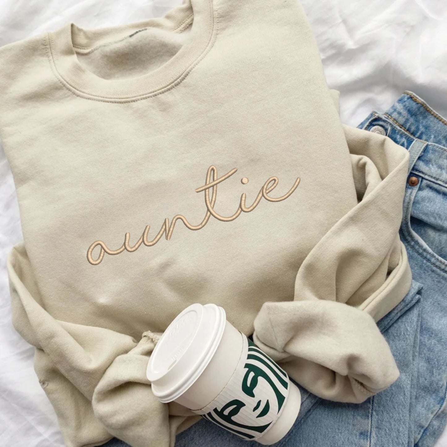 flat lay of a sand crewneck sweatshirt with embroidered auntie in lowercase script font and camel thread across the chest. The crewneck is styled with jeans and a starbucks cup