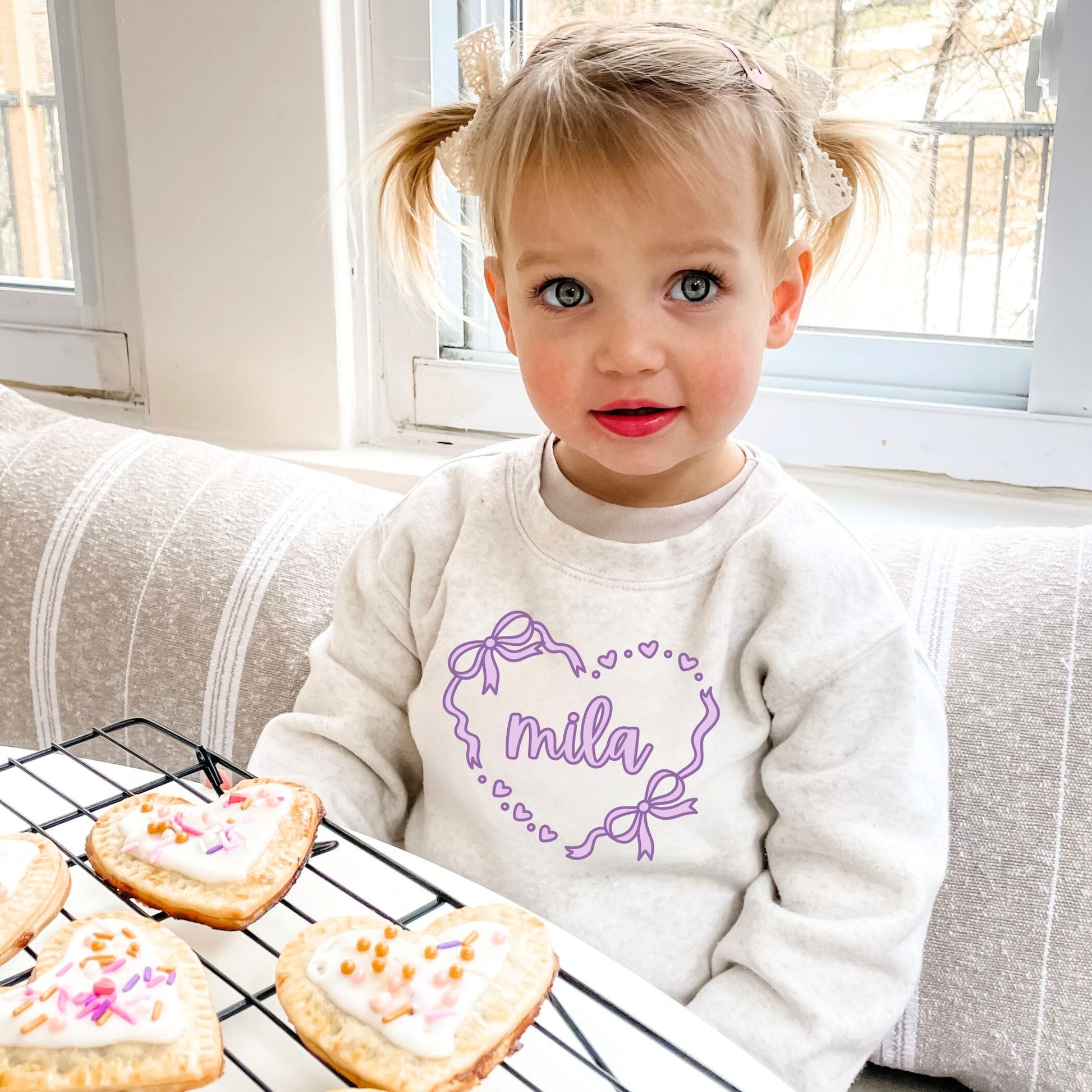 Child wearing a heather oatmeal sweatshirt with 'mila' heart design, sitting next to cookies on a cooling rack.