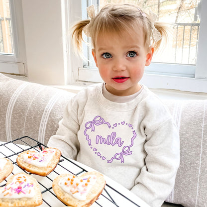 Child wearing a heather oatmeal sweatshirt with 'mila' heart design, sitting next to cookies on a cooling rack.