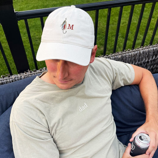 man sitting outside wearing a stone baseball cap with a fly fishing lure and single initial embroidered design on the center front