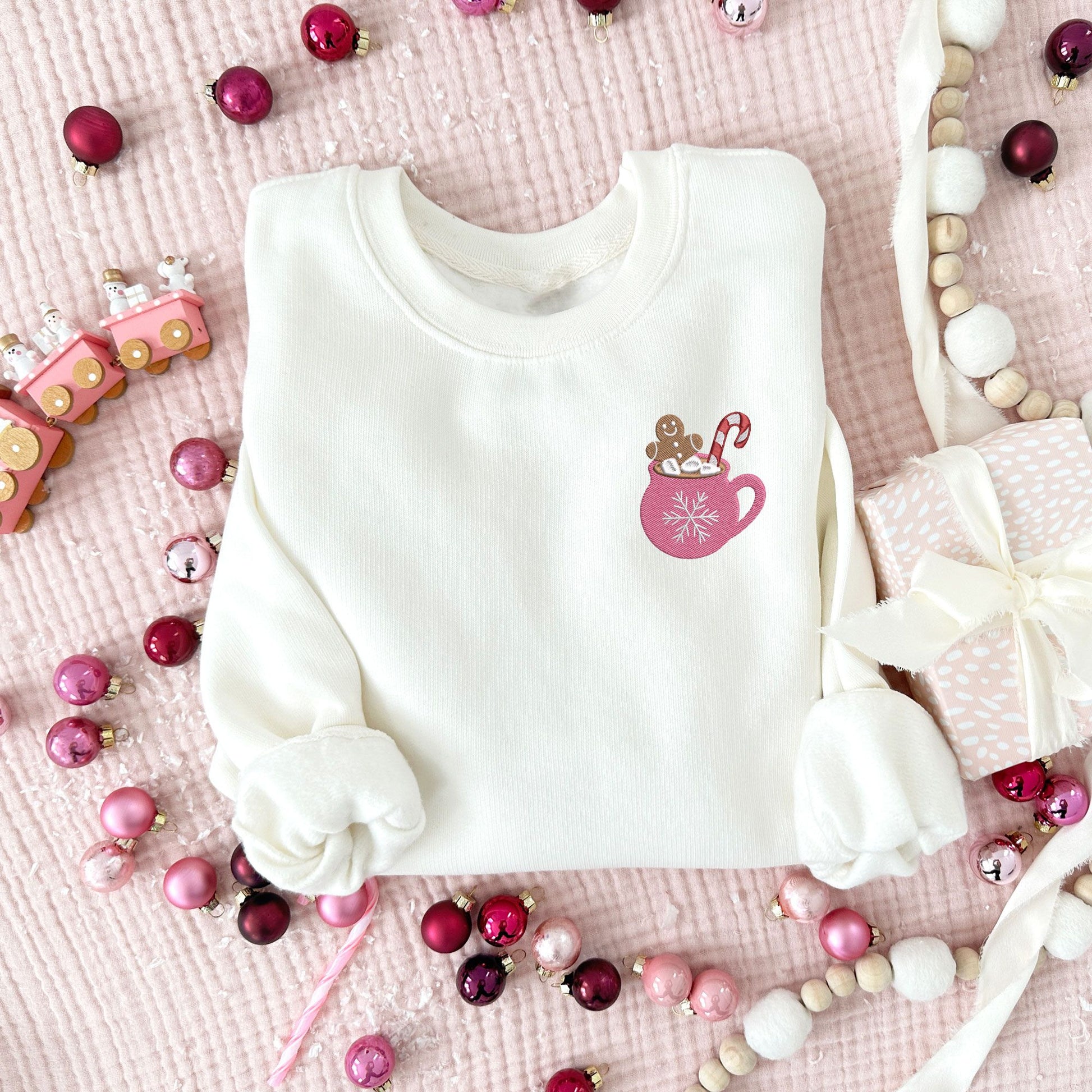 pink and festive Christmas flat lay photo of a sweet cream crewneck sweatshirt with embroidered pink mug with marshmallows a a ginger breadman and candy cane on the left chest.