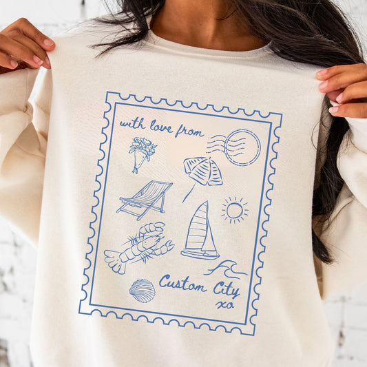 girl standing in front of brick wall wearing sweet cream sweatshirt with a printed design featuring a stamp outline with lobster, sailboat, folding chair, clam, waves, bouquet of flowers, umbrella, XO, and a space to add a custom city.