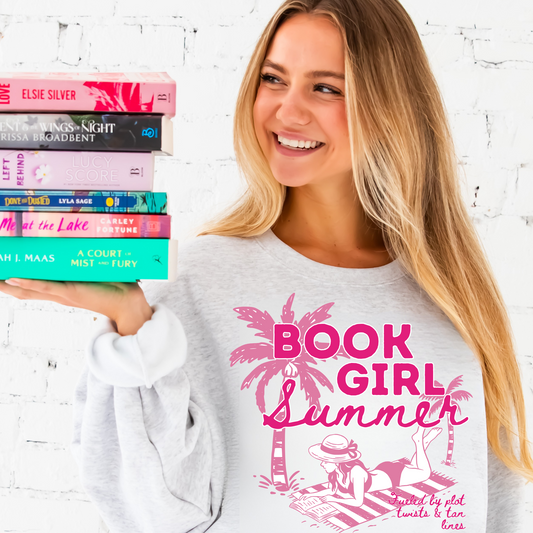 woman wearing ash sweatshirt with large pink printed Book Girl Summer design featuring palm trees, a girl laying out on a stripped beach towel reading a book and text that says book girl summer and plot twists and tan lines. She is holding a stack of books on her palm