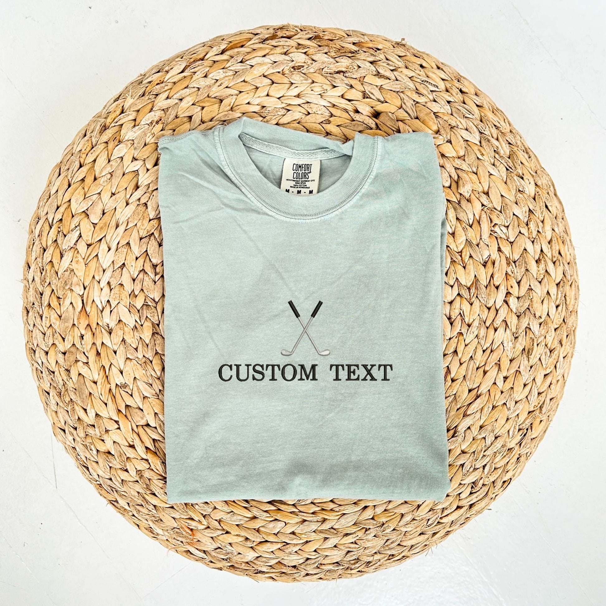 flat lay of bay comfort colors t-shirt with custom text and crossed golf club embroidery on the center chest