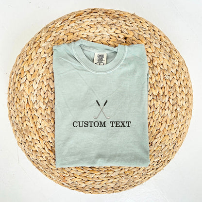 flat lay of bay comfort colors t-shirt with custom text and crossed golf club embroidery on the center chest