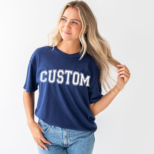 woman wearing a navy comfort colors t-shirt with a customizable printed design on the front center in a varsity style font