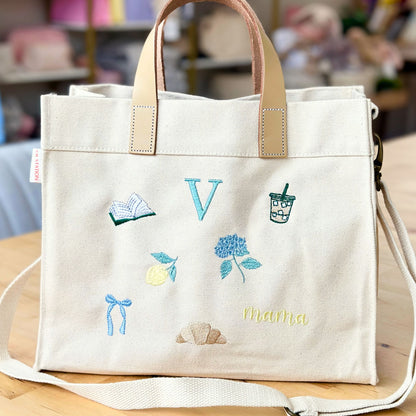 White tote bag with embroidered designs and 'Mama' text on a wooden surface.