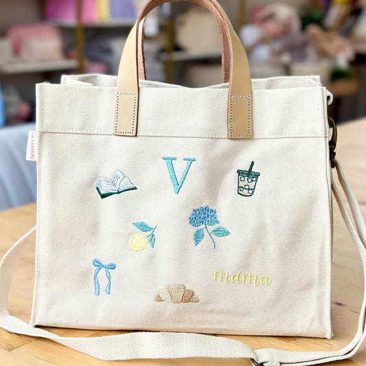 White tote bag with embroidered designs and 'Mama' text on a wooden surface.
