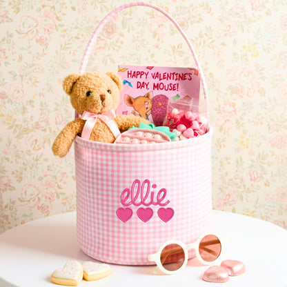 Pink gingham basket with script name and heart embroidery, stuffed with a teddy bear, candy, and 'Happy Valentine's Day, Mouse!' book on floral background