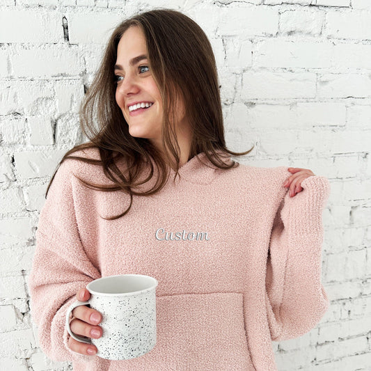 young woman wearing a pink hooded blanket with front kangaroo pocket in a soft microfiber fabric with a custom embroidered design on the front