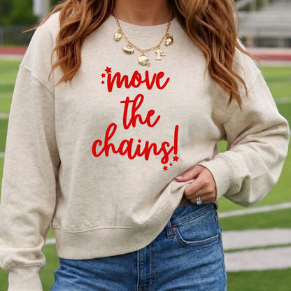 A woman wearing a cream-colored crewneck sweatshirt with the phrase 'move the chains!' printed in red across the chest, paired with blue jeans and gold necklaces, set against a blurred outdoor athletic field background.