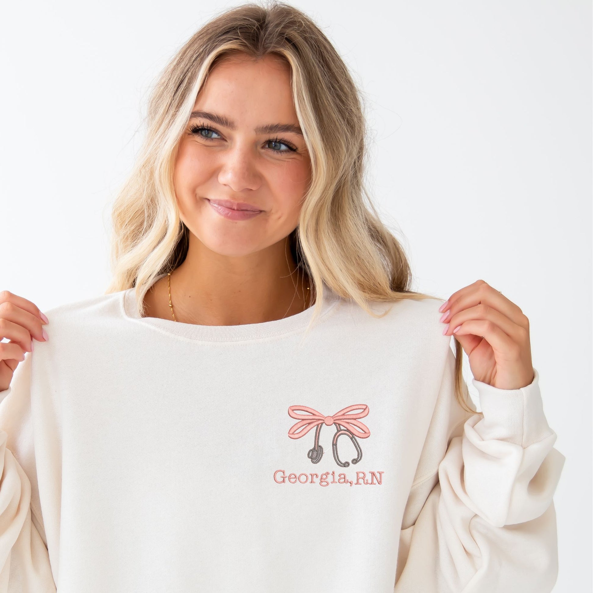 girl wearing a sweet cream crewneck sweatshirt with custom embroidered double loop bow stethoscope design and name on the left chest in coral pink and grey thread colors