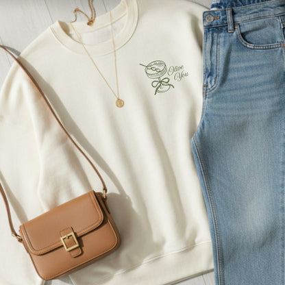 sweet cream sweatshirt  with olive you embroidered design on the left chest featuring a martini glass, bow, and olives and "olive you" text, paired with brown purse, and blue jeans 