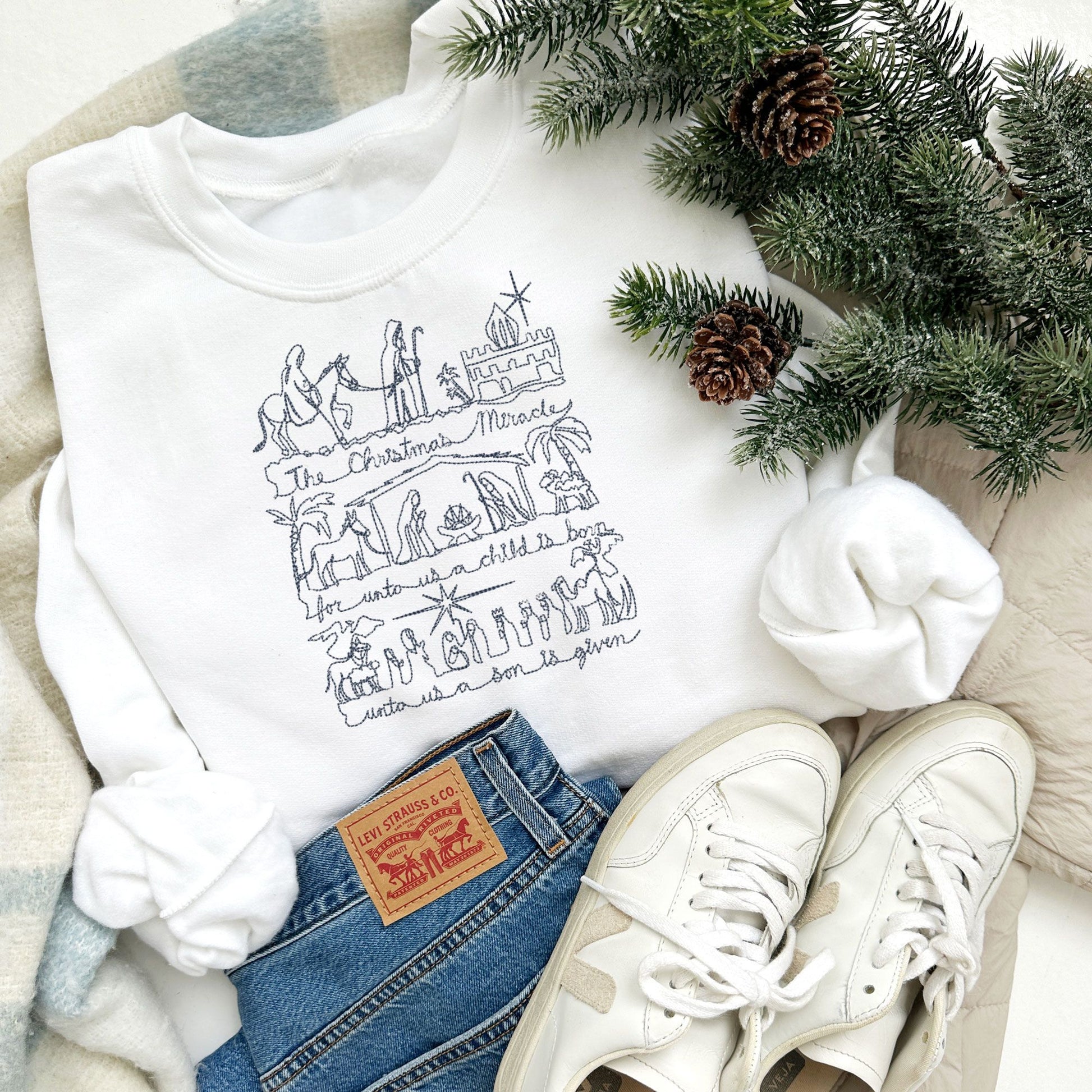 winter styled flat lay photo of a white crewneck sweatshirt with jeans and sneakers. Embroidered on the chest of the sweatshirt is a stitched drawing of the Christmas nativity in navy thread.