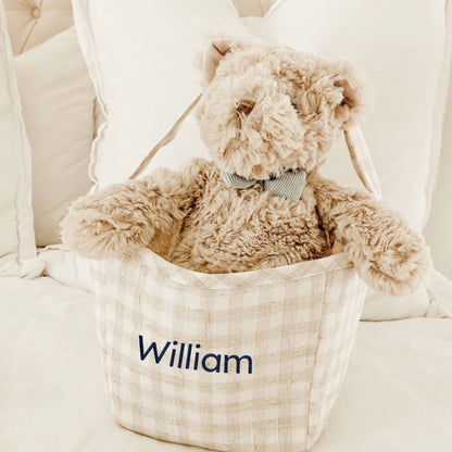 Teddy bear in a basket with 'William' embroidered on it, sitting on a white surface.