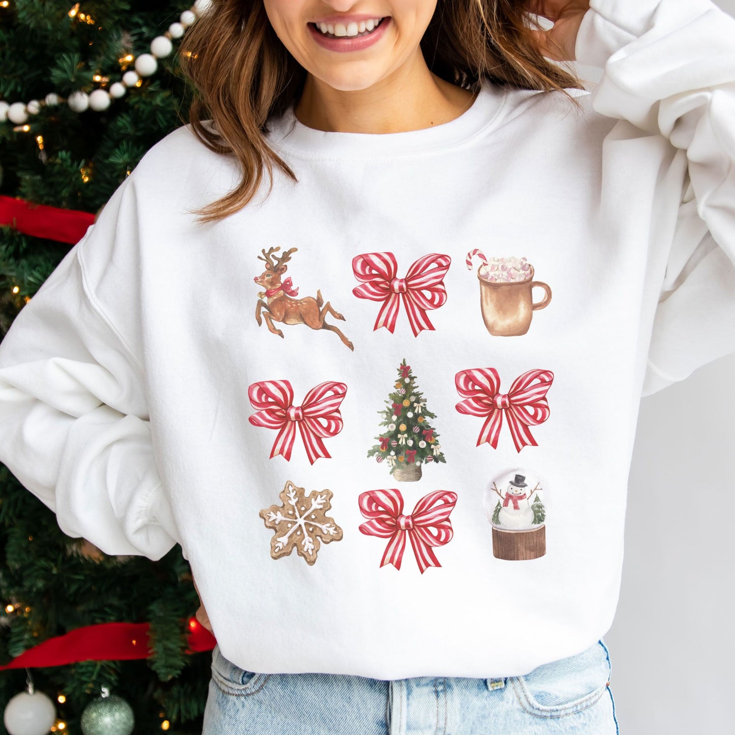 woman standing in front of a christmas tree wearing a white pullover with a retro reindeer, bow, cocoa, tree, cookie, and snow globe printed design