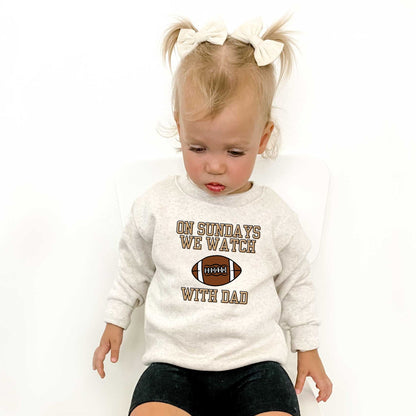 Little girl wearing a natural heather toddler crewneck sweatshirt with a custom on Sundays we watch football with dad print