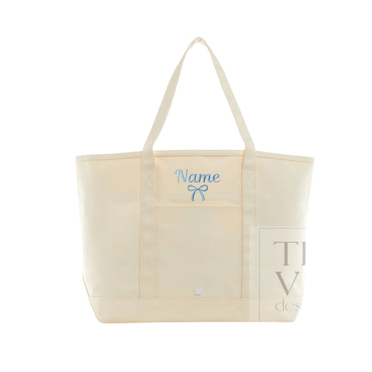 An off-white canvas tote bag with cream-colored handles, featuring a light blue embroidered bow and the word 'Name' on the front pocket, indicating personalization.