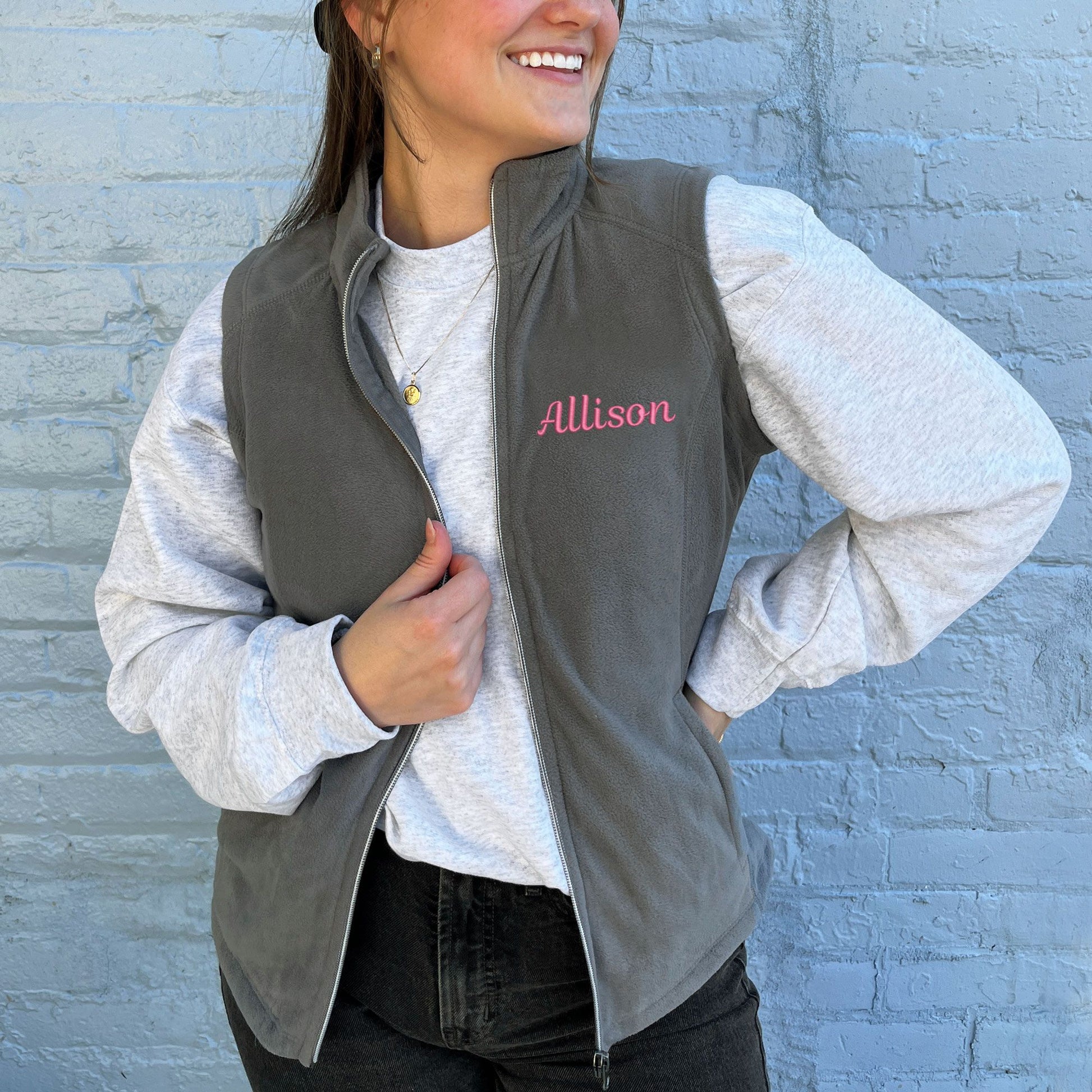 young woman wearing a lightweight grey fleece vest with name Allison  embroidered  on the left chest in pink thread