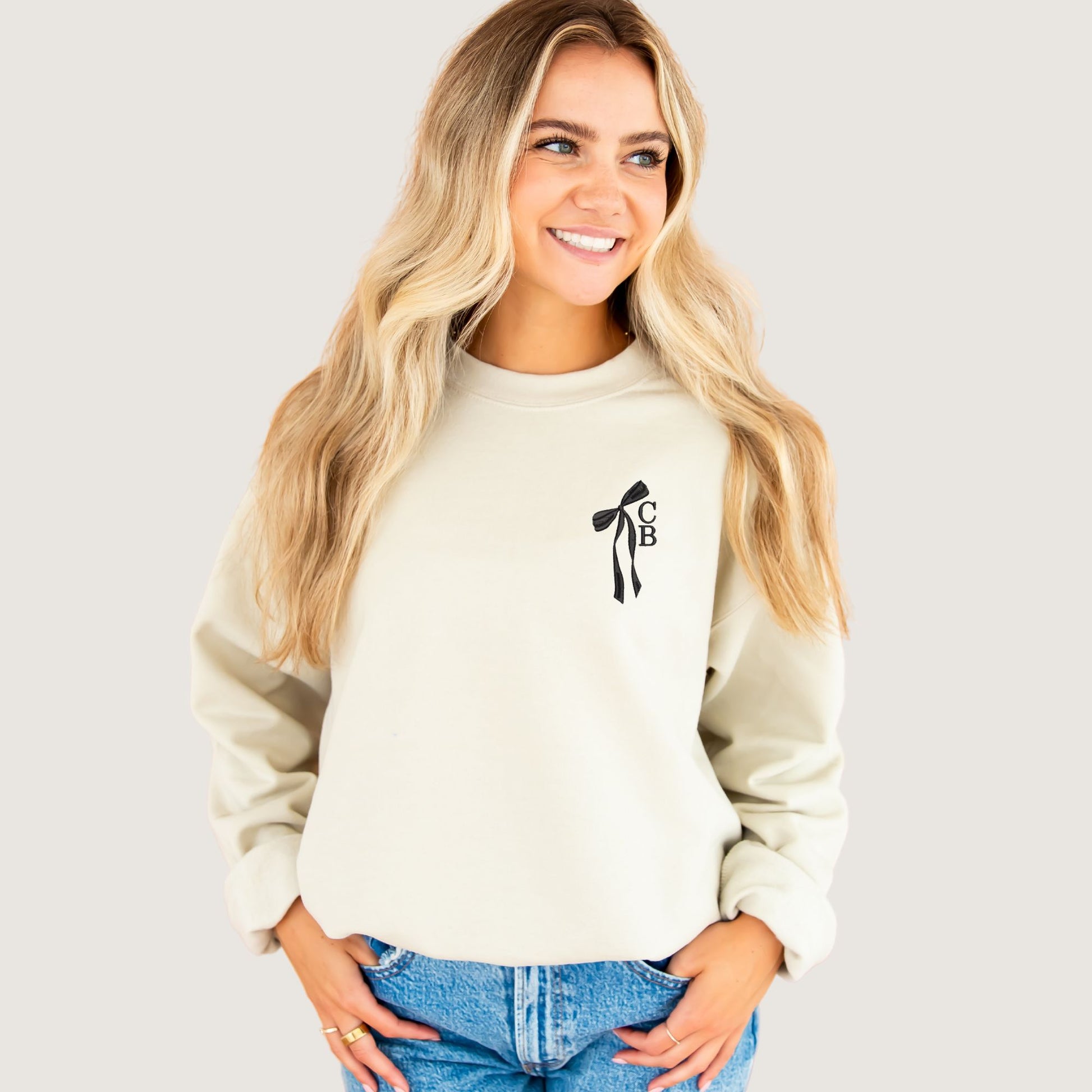 woman wearing a sand crewneck sweatshirt with custom ribbon bow and stacked initials embroidered on the left chest with black  thread