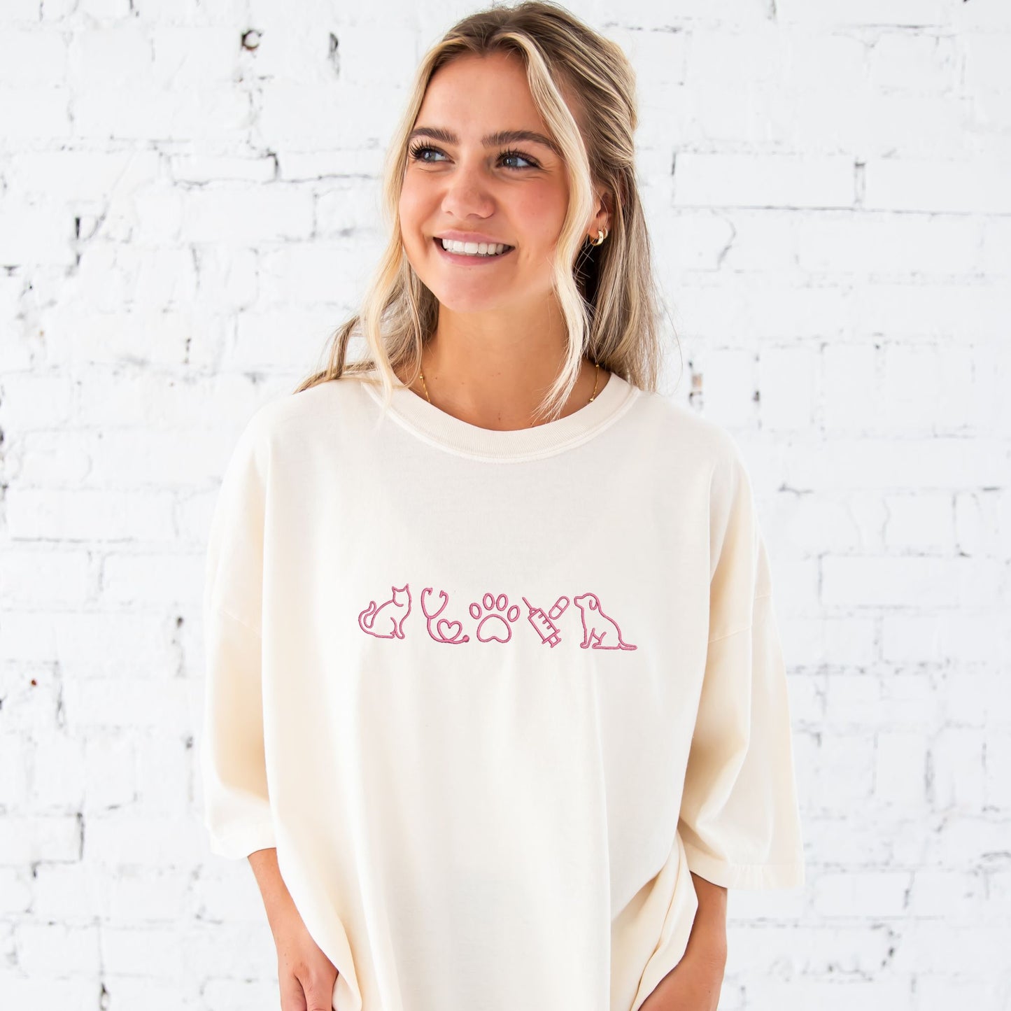 young woman wearing an ivory tee with embroidered veterinarian icons across the chest in pink thread.