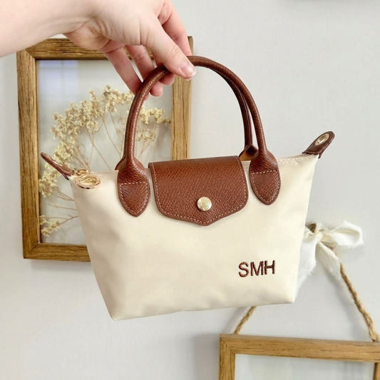 A hand holds a small cream canvas handbag with brown leather handles and trim. The bag has a flap front with a gold-tone snap button, a zip-top closure, and the letters 'SMH' embroidered on the front.